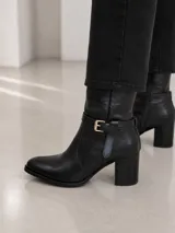 Bota de caña media confeccionada en cuero color marrón oscuro, con punta almendrada y taco cuadrado de 7 cm. Presenta una tira decorativa con hebilla metálica dorada en la zona del tobillo.