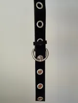 Cinturón de símil gamuza color tostado, con diseño de ojales metálicos plateados a lo largo de toda la pieza y hebilla redonda metálica.