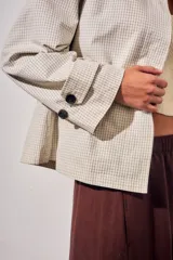 Trench corto de corte oversize con estampado de cuadros finos en tonos beige y verde. Presenta cuello camisero en contraste de color marrón oscuro, cierre frontal con botones y mangas largas con detalle de trabas en los puños.