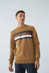 Buzo color caramelo de felpa con cuello redondo, mangas largas, puños y cintura acanalados. Presenta una franja horizontal en el pecho con el logo de la marca Mistral y la inscripción "NETHERLAND EST 76".