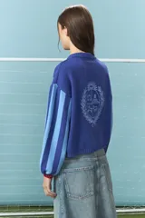 Cardigan de algodón color azul con mangas largas de rayas verticales anchas en tonos azul y celeste, puños en rojo, y bordado en la espalda con diseño de escudo nacional.