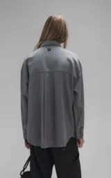 Camisa de manga larga en tejido twill color gris melange, con corte oversize. Presenta cuello clásico, cierre frontal con botones y tres bolsillos delanteros, uno de ellos oculto.