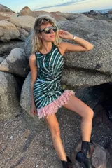 Conjunto de dos piezas compuesto por un top sin mangas y una falda corta, ambos confeccionados en tela de lentejuelas con diseño de rayas diagonales en tonos verde y plateado. La falda presenta un borde inferior decorado con plumas rosadas.