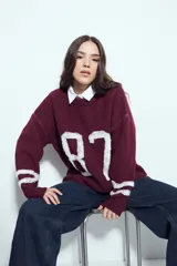 Sweater de tejido de punto color blanco con estampado numérico '87' en negro en el frente y detalles de rayas horizontales en las mangas. Presenta cuello redondo y un calce relajado.