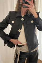 Chaqueta corta de cuero negro con cuello clásico, cierre frontal con botones grandes y bolsillos de parche en el pecho.