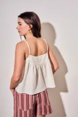 Blusa color off white de algodón peinado, sin mangas, con tirantes finos, escote en V con detalles de encaje y bordados, y pliegues decorativos en los laterales.