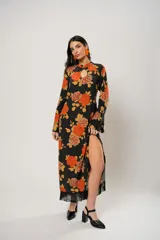 Vestido largo negro con estampado de flores naranjas y amarillas, cuello mao con abertura en forma de gota, mangas largas acampanadas con botones en los puños, tajo lateral y flecos en el ruedo.