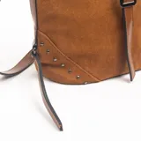 Cartera de gamuza color marrón con diseño estructurado, correa ajustable y detalle de tira central con hebilla metálica. Presenta apliques de tachas metálicas en las esquinas inferiores y correa larga extraíble.
