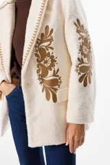 Blazer de textura suave en color crudo, con solapas clásicas y bordados florales en tonos tierra en el frente y las mangas. Presenta detalles de costura decorativa en los bordes de las solapas.