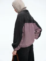 Campera de estilo casual con diseño bicolor, combinando paneles en color gris oscuro y malva. Presenta cuello alto con cierre frontal de botones a presión, mangas largas con puños elásticos y ruedo ajustable con cordón.