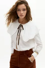 Camisa blanca de lino con cuello Peter Pan grande y ondulado, mangas largas y cierre frontal con botones de nácar. Incluye lazo decorativo de terciopelo marrón en el cuello.