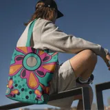Bolso de tela tipo tote bag con estampado de flores de estilo retro en tonos turquesa, naranja, rosa y negro. Tiene asas de color turquesa.