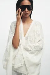Blusa tipo capa de algodón con escote en pico y mangas largas amplias. Presenta bordados florales a tono en toda la prenda, bajo asimétrico y cierre frontal con botones.