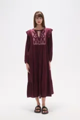 Vestido midi holgado de viscosa color vino tinto, con mangas largas y volados en los hombros. Presenta un canesú con bordado floral en tonos rosados y cuello con abertura en forma de lágrima y lazo para atar.