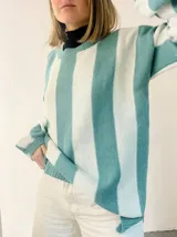 Sweater de punto con diseño de rayas verticales en tonos azul y gris, con sutil acabado brillante. Presenta cuello redondo, mangas largas con puños ajustados y corte amplio con aberturas laterales en el ruedo.