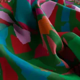 Pañuelo cuadrado de seda con estampado abstracto multicolor en tonos verde, rosa, rojo, naranja, azul y violeta. Tiene un borde liso color fucsia y un borde interno con diseño de soga.