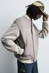 Cazadora estilo varsity o bomber de hombre, color gris oscuro con efecto ante, mangas en tono gris claro y puños, cuello y bajo con rib elástico a rayas blancas y grises. Presenta un parche con la letra 'R' en el pecho derecho y un bordado con texto en el pecho izquierdo.