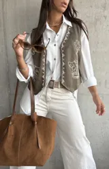 Chaleco de gamuza en color verde oliva, con diseño de bordados florales en hilo blanco y detalles de tachas metálicas. Presenta un corte clásico con cierre frontal de botones.