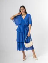 Vestido midi de seda italiana color azul, con escote en V, mangas murciélago amplias, falda de tres volados y cintura elastizada con lazo ajustable al tono y hebilla circular dorada.