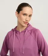 Campera deportiva de mujer con cierre frontal, capucha ajustable con cordones, mangas largas con puños y bolsillo canguro. Confeccionada en una mezcla de modal, poliéster y elastano, presenta un diseño funcional con sisa raglán y recorte en la espalda.