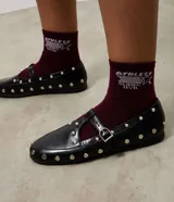 Zapato tipo salomé de color negro con acabado liso, puntera redondeada y suela plana. Presenta una tira central con hebilla metálica ajustable y está decorado con tachuelas metálicas plateadas distribuidas por toda la superficie.