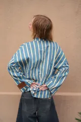 Camisa de denim azul con cuello mao, diseño holgado, mangas largas y cierre frontal con botones metálicos. Cuenta con costuras decorativas pespunteadas en el delantero.
