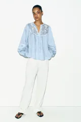 Blusa de rayas verticales blancas y celestes, con cuello redondo y escote en pico. Presenta detalles de bordados perforados y puntillas en el pecho y las mangas. Tiene mangas largas abullonadas con puños elásticos y cierre frontal con botones forrados.