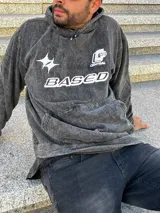 Buzo gris nevado con capucha y bolsillo tipo canguro. Estampado frontal con la palabra "BASED" y un logo.