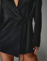 Vestido tipo blazer negro de corte entallado, con solapas clásicas, mangas largas y cierre frontal cruzado con lazo ajustable en la cintura.