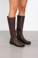 Botas de caña media confeccionadas en cuero vacuno, con diseño de punta redondeada y suela plana con pequeño taco cuadrado. Presentan costuras verticales decorativas a lo largo de la caña.