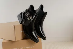 Botineta de cuero charol negro con acabado brillante, diseño de punta fina y taco cuadrado. Presenta un panel elástico lateral y una correa decorativa con hebilla metálica plateada.