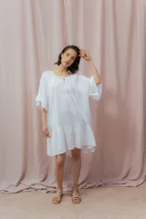 Vestido corto blanco de lino, con diseño suelto y holgado, mangas amplias hasta los codos, escote redondo con abertura en V y lazo decorativo, y un volante en la parte inferior.