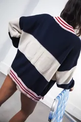 Cardigan oversized de punto grueso, color azul marino y blanco con franjas rojas y rosadas en el cuello, puños y solapa. Tiene botones grandes marrones y el logo de Anthea bordado en el pecho.