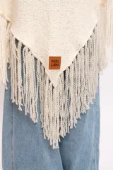 Poncho triangular de lana merino color marfil, confeccionado artesanalmente en telar manual con textura rústica y delicados flecos colgantes en los bordes.