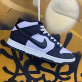 Championes Nike Dunk Mid con diseño bicolor en blanco y negro. Confeccionados en lona resistente, presentan el icónico swoosh de Nike en los laterales, cierre con cordones y suela de goma con efecto moteado.