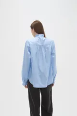 Camisa celeste de manga larga con cuello alto y volados, detalles de frunces en hombros y puños, y cierre frontal con botones.