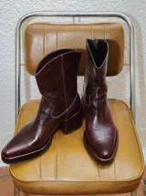Bota estilo tejano de cuero color bordó con acabado brillante, taco cuadrado y punta cuadrada. Presenta costuras decorativas en el empeine y caña media.