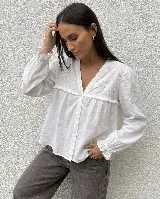 Camisa blanca de corte amplio con escote en V, canesú delantero con bordado floral y detalle de volado pequeño. Presenta mangas largas con puños elastizados y cierre frontal de botones.