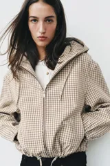 Campera estilo cazadora con capucha ajustable con cordones, cierre frontal de cremallera, mangas largas con puños elásticos, bolsillos delanteros de doble vivo y bajo ajustable con cordones. Presenta un diseño de cuadros en tonos beige y marrón.