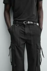 Pantalón cargo de corte relajado en color negro con efecto lavado. Presenta bolsillos laterales tipo cargo con solapas y cintas ajustables, bolsillos frontales y traseros, y detalles de argollas metálicas. Cierre frontal con cremallera y botón.