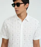 Camisa de manga corta para hombre, confeccionada en algodón color blanco con diseño de bordado de flores y perforaciones geométricas. Presenta cuello inglés y cierre frontal con botones.