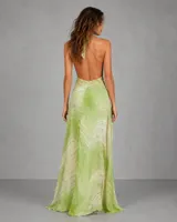 Vestido largo asimétrico de estilo etéreo, con escote halter cruzado y espalda descubierta. Presenta un diseño corto en la parte frontal y una cola larga en la parte posterior, confeccionado en una tela ligera con estampado abstracto en tonos verdes.