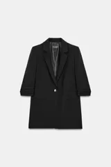 Blazer negro con cuello solapa con muescas, hombreras y forro interior a tono. Presenta manga por debajo del codo con detalle de drapeado y falsos bolsillos de vivo en el delantero, con cierre frontal de un solo botón dorado.