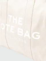 Bolso tipo tote de Marc Jacobs, confeccionado en lona resistente de color negro. Presenta el nombre del modelo estampado en blanco en la parte frontal y el logo de la marca debajo. Cuenta con doble asa superior para llevar al hombro o en la mano.