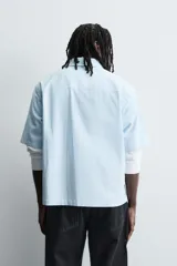 Camisa de corte relajado confeccionada en tejido de mezcla de algodón, con estampado de rayas verticales en tonos azul y blanco. Presenta cuello solapa, manga corta y cierre frontal con botones. Incluye un detalle bordado a contraste en el pecho.
