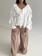 Blusa blanca de corte holgado con escote en V, decorada con volados frontales y en las mangas. Presenta mangas largas con puños elásticos y cierre frontal con botones.
