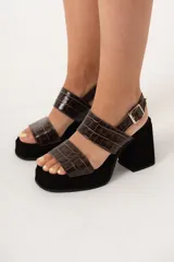 Sandalias de tacón alto con plataforma, color negro y marrón con estampado de cocodrilo. Tienen dos tiras anchas sobre el empeine y una tira con hebilla alrededor del tobillo. El tacón es ancho y cuadrado.