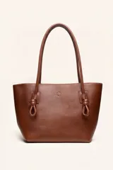 Bolso tipo tote color marrón de cuero suave, con doble asa larga decorada con nudos en los extremos inferiores y detalle de logo grabado en el frente.