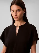 Blusa Calvin Klein de manga corta en color negro, confeccionada en tejido fluido de crêpe de chine. Cuenta con un diseño de cuello redondo con escote en V y una barra metálica decorativa en el centro del cuello.