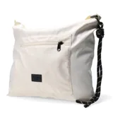 Cartera tipo bandolera de color beige, confeccionada en material sintético ligero. Presenta un bolsillo frontal con cierre, un parche decorativo con el logo de la marca y una correa ajustable de cordón técnico negro con detalles blancos.